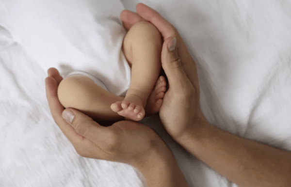 Close-up of a parent's hands gently cradling a newborn baby's tiny feet and legs on white bedding.
