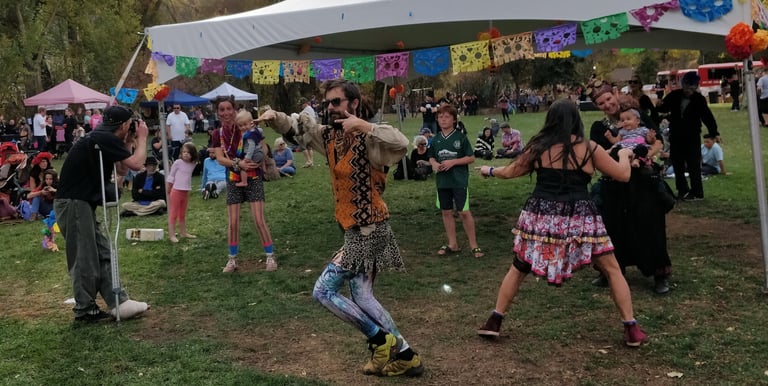 Dancers in eclectic costumes perform at an outdoor community festival under a white canopy.