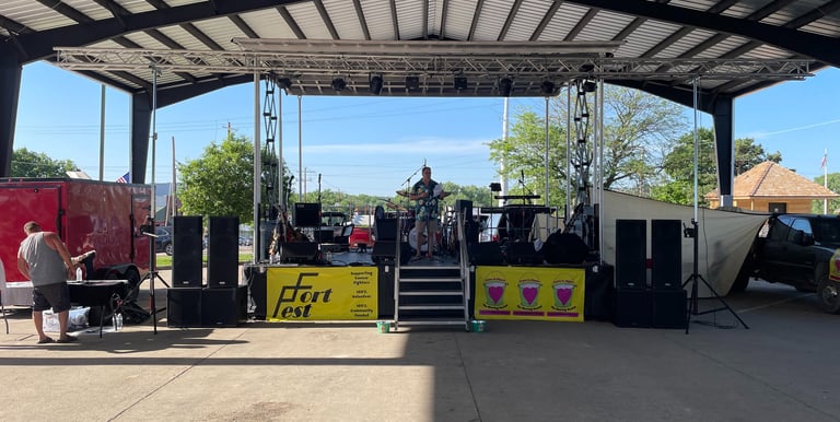 A musician performs on an outdoor stage at Fort Fest under a large metal pavilion roof with professional audio equipment.