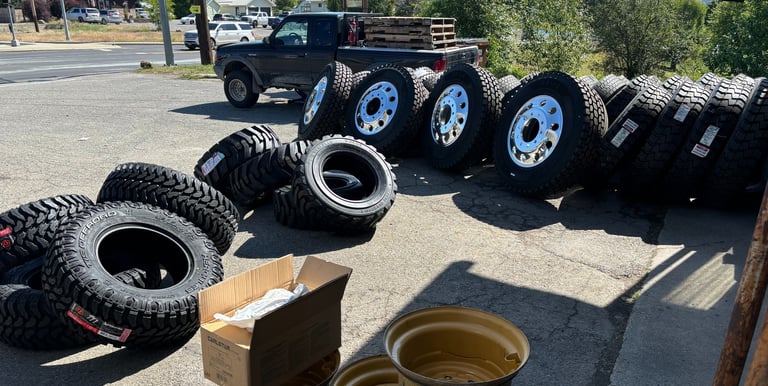 A collection of heavy-duty truck tires and chrome rims displayed on asphalt at a tire service shop.