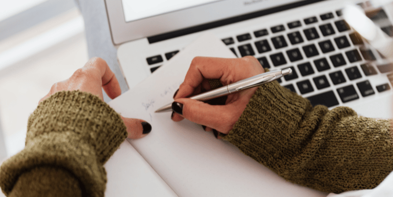 Hand holding a notebook page and writing with laptop in the background