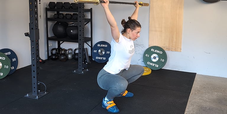 A woman performing an overhead barbell squat in a home garage gym with weight plates.