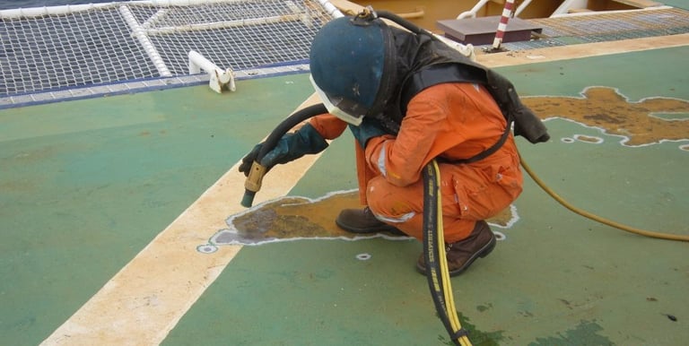 A worker in safety gear sandblasting a metal ship deck for corrosion protection and maintenance.