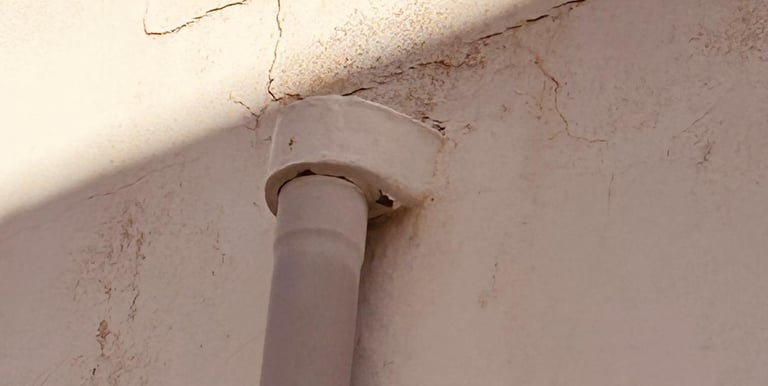A vertical drainage pipe attached to a white exterior wall with visible cracks and shadows.