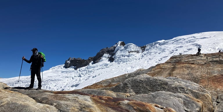 A hiker with trekking poles stands on a rocky ridge overlooking a snow-covered mountain glacier.