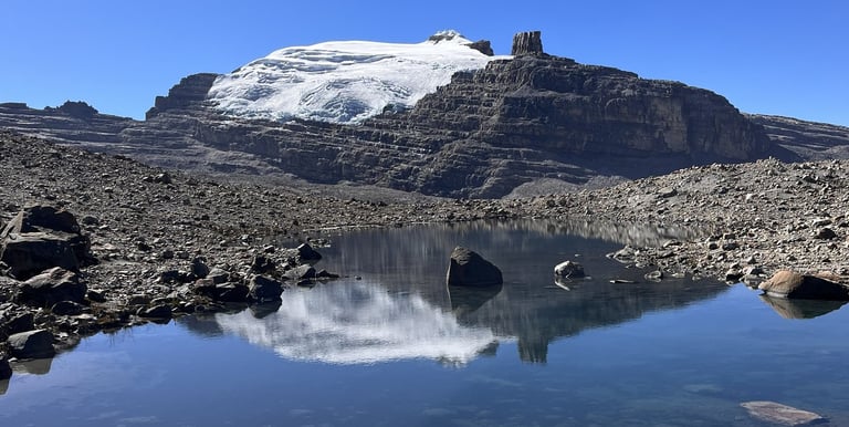 Snow-capped mountain peak reflected in a clear alpine glacial lake under a bright blue sky.
