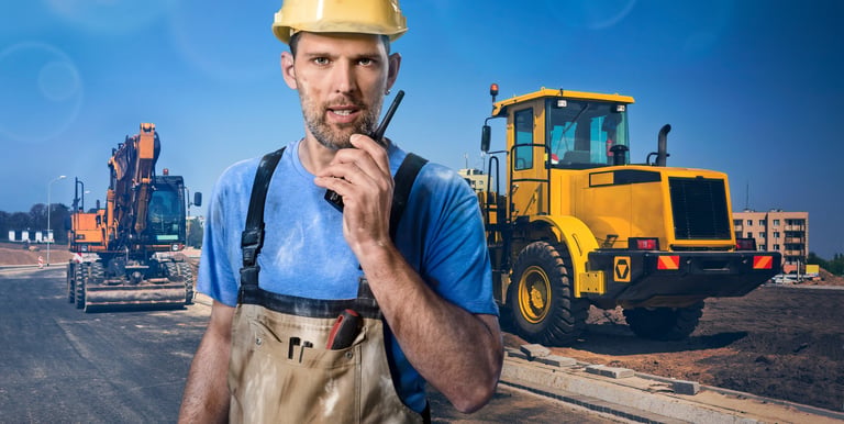 A construction worker in a yellow hard hat using a walkie-talkie at a roadwork site with heavy machinery.