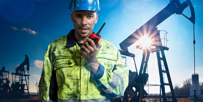 Oil field worker in high-visibility gear using a walkie-talkie near a pump jack at sunset.