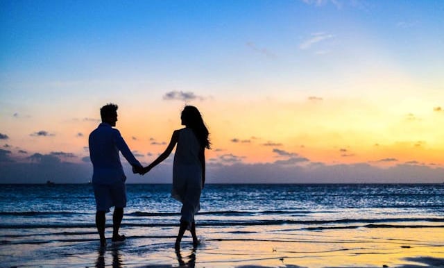 A romantic couple silhouette holding hands on a tropical beach during a colorful sunset.