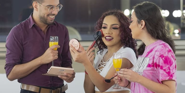 Maquilladora profesional aplica rubor a una mujer sonriente mientras sus amigos disfrutan de bebidas