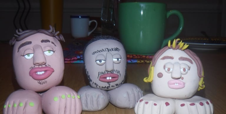 Three handmade clay head sculptures with feet on a wooden table near a green coffee mug.