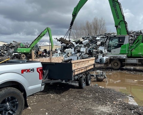 a truck and trailer being unloaded at metal recycling facility by large green tractor with a magnet