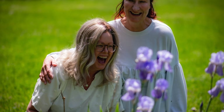 Two laughing women in white tops enjoying blooming purple iris flowers in a sunny green garden.