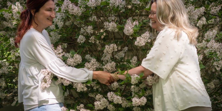 Two women shaking hands outdoors in front of a blooming white lilac bush garden.