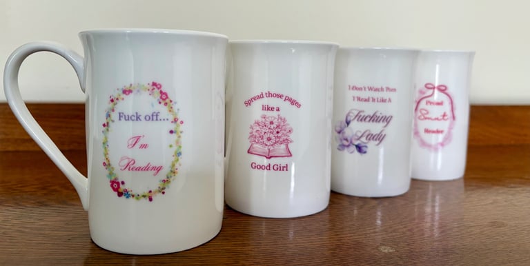 Four white bone china mugs with pink spicy romance book trope quotes on a wooden table.