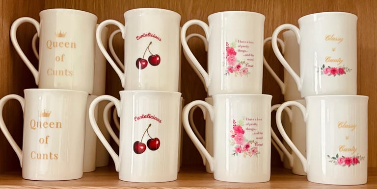 White bone china mugs with floral designs and bold profane slogans on a wooden shelf.