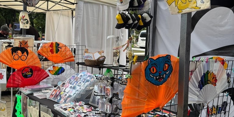 Puesto de venta artesanal en feria creativa con abanicos pintados a mano, stickers de anime y pop