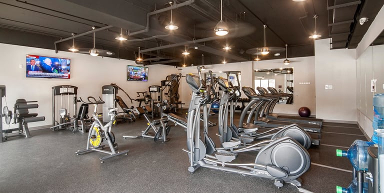 Modern apartment gym featuring elliptical machines, treadmills, stationary bikes, and strength training equipment.