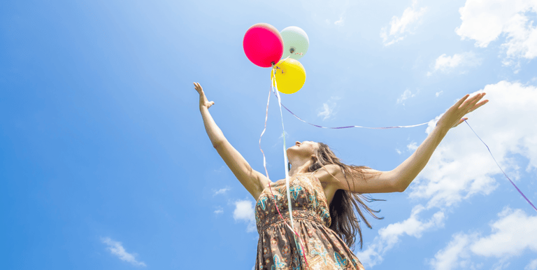 Mujer joven en un espacio abierto que muestra un cielo azul, lanzando tres globos al aire, feliz.