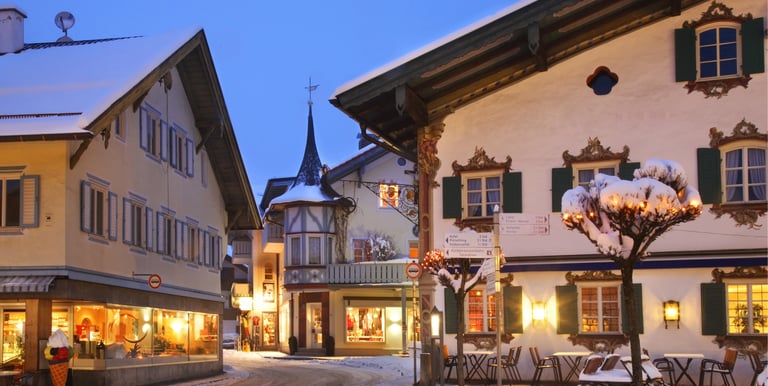 Snowy Bavarian village street at dusk with illuminated traditional alpine chalets and shops.