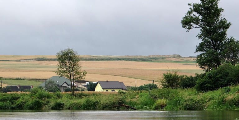 Widok ze śrdoka rzek blisko wody. Rzeka zakręca a na zakręcie w oddali domy. Dalej domy i pole
