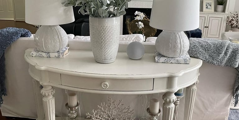 Painted sofa table in off white with light distressing with two lamps and a vase with greenery. 