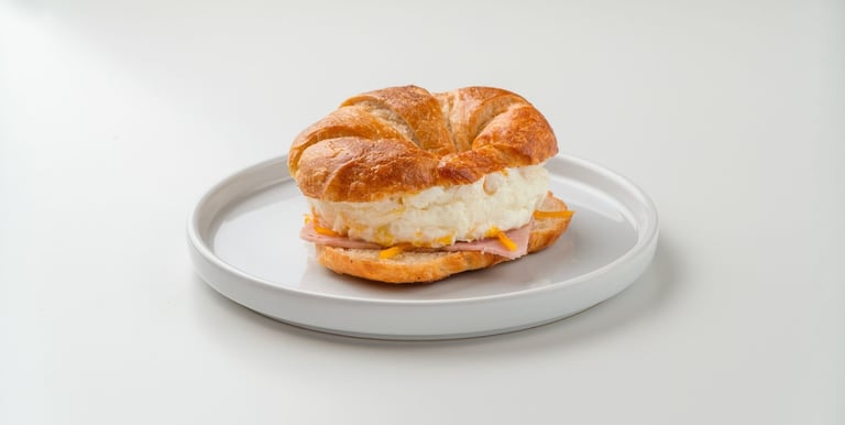 Breakfast croissant sandwich with fluffy eggs, melted cheese, and ham on a white plate.