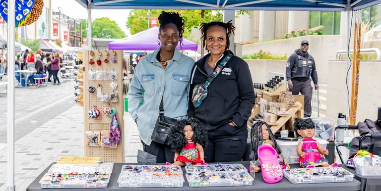 Two women smiling at an outdoor Afro Fiesta Festival market stall selling handmade accessories and dolls.