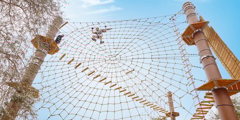 A person climbs a giant spider web rope course at an outdoor adventure park under a clear blue sky.