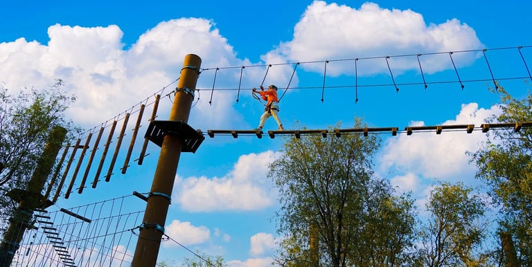 A person navigates a high ropes adventure course with wooden bridges and cables against a blue sky.