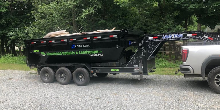 A black Riverbend Rolloffs and Landscape dump trailer loaded with debris on a gravel driveway.