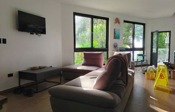 Modern bright living room with a large plush sectional sofa, wall-mounted TV, and floor-to-ceiling windows overlooking lush
