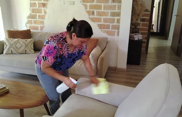 Professional housekeeper cleaning a white fabric armchair with a spray bottle and cloth in a living room.