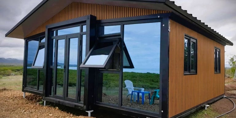 Modern prefab container home with wood cladding and large reflective glass windows in a rural landscape.