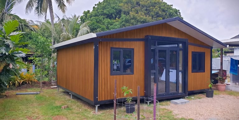 Modern wooden prefab tiny home with black trim nestled in a tropical garden landscape.