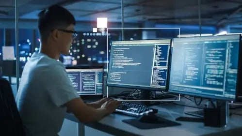 muestra una chica sentada mirando 2 monitores de PC en una sala computarizada