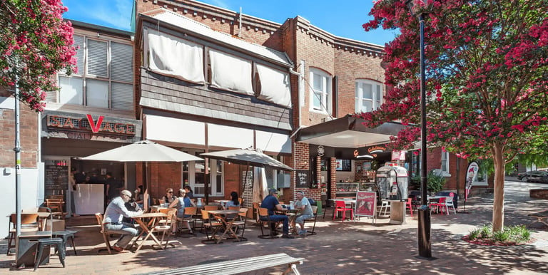 People dining at an outdoor sidewalk cafe with red brick buildings and blooming pink trees.