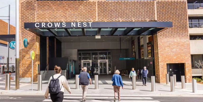 Commuters crossing the street toward the modern brick entrance of Crows Nest Metro station.