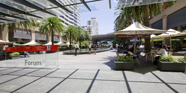 Sunny modern outdoor Forum Plaza with palm trees, glass awnings, and cafe seating in an urban shopping area.