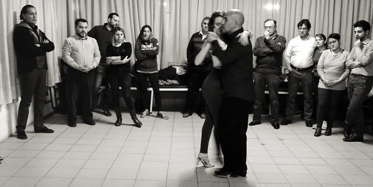Una pareja bailando tango argentino en un estudio de baile, con alumnos observando.