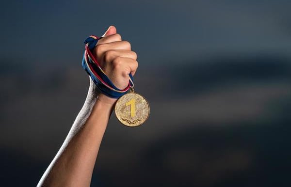 Atleta com o braço ergudi segurando uma medalha de ouro com 1º lugar