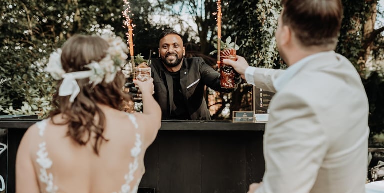 A smiling wedding bartender serves sparkling cocktails to a bride and groom at an outdoor garden bar.