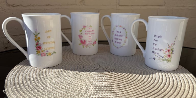 Four white bone china mugs with floral designs and funny, profane quotes on a woven placemat.