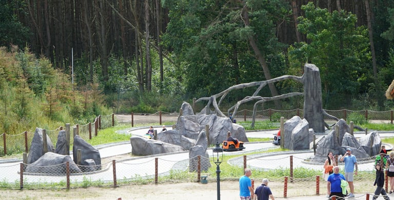 Families enjoy an outdoor go-kart track with stone obstacles and a forest backdrop at a theme park.