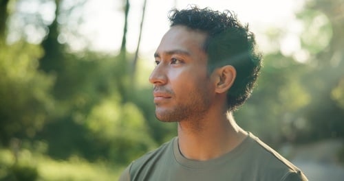 A profile of a young man with curly hair standing in a sunlit park with lens flare and greenery.