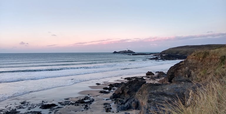 Scenic coastal view of Godrevy Lighthouse at sunset from a rocky beach in Cornwall, England.