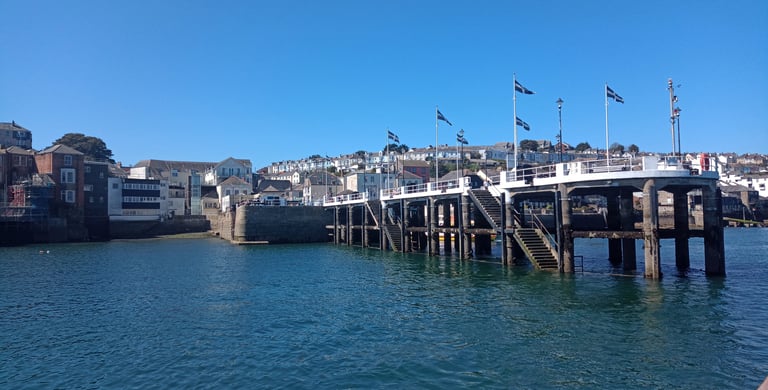 Concrete pier with flags in Falmouth, Cornwall, under a clear blue sky with coastal town houses.