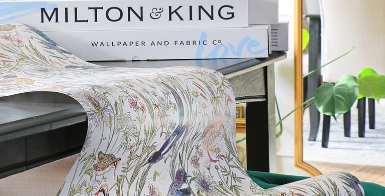 Milton and King floral bird wallpaper roll draped over a green velvet ottoman in a bright room.