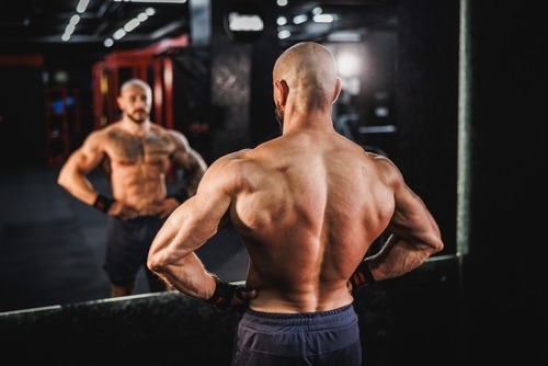 Muscular bodybuilder flexing back muscles and lats in front of a gym mirror.