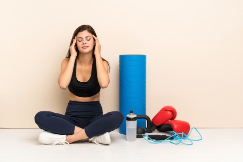 A woman in fitness wear sitting with closed eyes next to a yoga mat, boxing gloves, and workout gear.